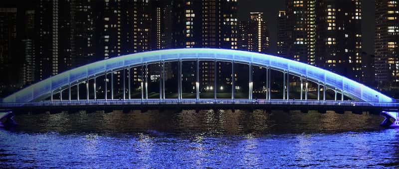 Imagen de ejemplo de un puente reflejado sobre el agua de noche con edificios en el fondo